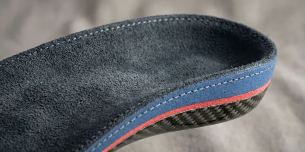 A high-detail macro studio photograph of a custom orthotic insole. The composition focuses on the heel profile, showcasing a dark gray suede-textured top with precision white stitching. Layers of blue and red high-density foam are visible above a glossy, woven carbon fiber base. The lighting is soft and diffused, highlighting the technical craftsmanship and premium materials against a neutral, out-of-focus background.