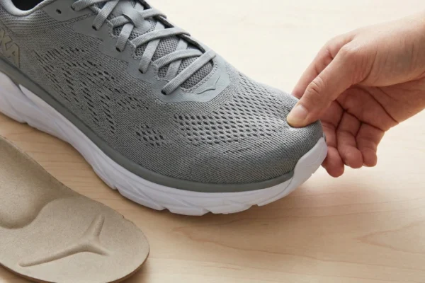 A detailed close-up shot on a light wooden surface under soft, natural light. A hand is seen pressing the wide toe area of a grey mesh athletic shoe to demonstrate its flexibility and roominess. In the lower-left corner, a tan-colored custom orthotic insole lies flat, showing its anatomical contours and support structure. The composition focuses on the texture of the breathable fabric and the practical application of corrective footwear.