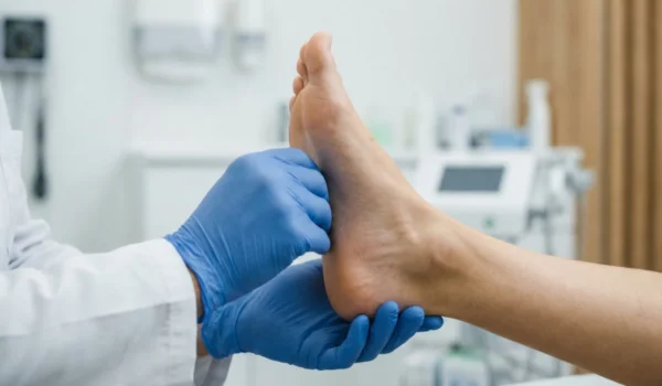 A close-up, high-angle shot featuring soft, diffused clinical lighting. A practitioner in a crisp white lab coat and textured blue nitrile gloves performs a manual examination of a patient’s bare foot. The composition is tightly focused on the tactile interaction, with a blurred background showing a modern medical monitor and clean, minimalist clinic interiors.