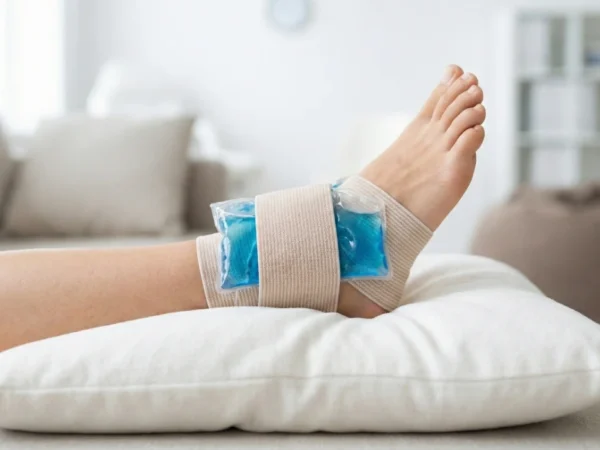 A photograph of a person's ankle elevated on a soft, white pillow. A clear, reusable gel ice pack, showing blue gel inside, is secured firmly around the ankle with a beige elastic compression bandage. The person is resting comfortably on a light gray couch in a brightly lit, modern living room with blurred furniture and a window in the background.