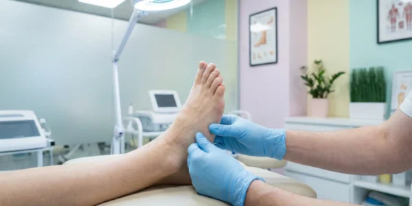 A crisp, brightly lit clinical photograph showing a podiatrist’s hands in blue nitrile gloves performing a manual examination of a patient’s foot. The composition is clean and professional, set in a modern medical office with pastel-colored walls, a bright examination lamp, and professional medical equipment visible in the soft-focus background.