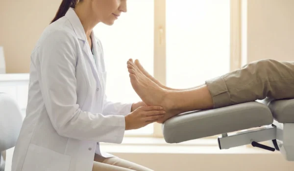 A female podiatrist in a white coat carefully performs a manual examination of a patient's foot in a clinic setting.
