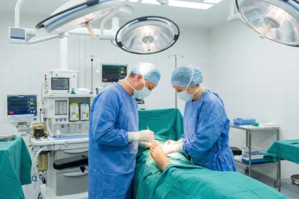 A wide-angle shot of a sterile operating room illuminated by three large, bright surgical LED lamps. Two surgeons in blue scrubs, masks, and caps lean over a patient's foot draped in green medical fabric. The composition is centered on the surgical site, with blurred medical monitors and anesthesia equipment in the background. The lighting is cold, clinical, and high-intensity.
