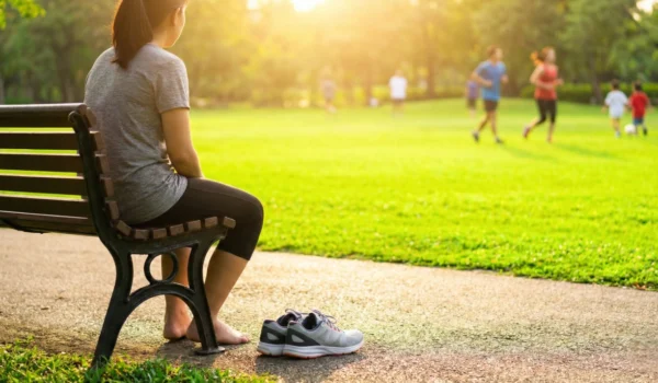 A barefoot woman sits alone on a park bench, watching others exercise in the distance, her sneakers placed beside her.