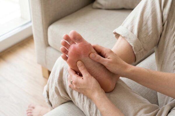 A close-up, elevated photograph shows a person seated on a textured beige sofa, wearing light-colored linen pants, looking down at their raised foot. Bright, natural daylight from the left clearly illuminates their hands gently holding and applying pressure to the bare sole of the foot, which is turn towards the viewer. The metatarsal heads (ball of the foot) exhibit distinct red, inflamed areas. A wedding ring is visible on the finger of the left hand. The shallow depth of field keeps the focus sharp on the interaction between the hands and the painful-looking foot, with the blurred background of the sofa and a wooden floor providing context.