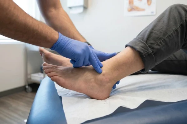 A side-view close-up shot with soft natural lighting illuminating a patient's bare foot resting on a white-papered blue medical table. A practitioner in blue latex gloves is seen gently pressing fingers against the ankle joint. The background shows a blurry medical office setting with neutral colors and soft shadows.